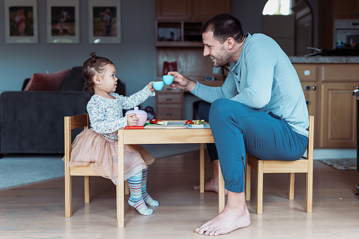 Profile view of a dad and his toddler daughter sitting at a child size table and doing a celebratory toast with toy teacups while playing together at home.