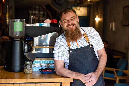 Portrait of urban mid-adult Caucasian man, an small business owner, wearing apron, while standing at coffee shop