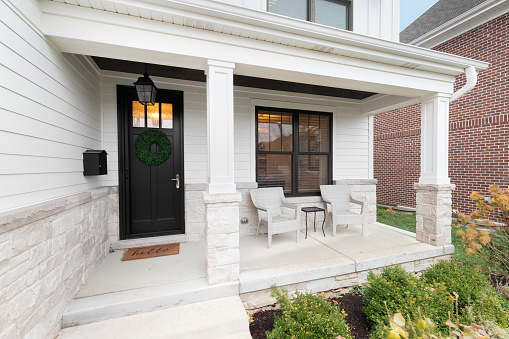Oak Park, IL, USA - November 15, 2020: A new, white modern farmhouse with a dark wood door with windows, white pillars, a stone floor, and patio furniture.