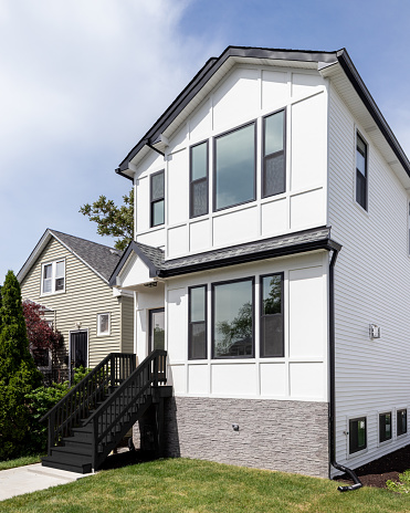 Oak Park, IL, USA - April 25, 2021: A new, white modern farmhouse with a dark shingled roof and black window frames. The bottom of the house has a light rock siding and black stairs.