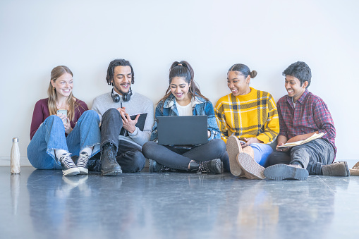 A small group of University students sit on the floor together as they casually work on an assignment together.  They have one laptop open that they are each sharing and other students are holding notebooks.