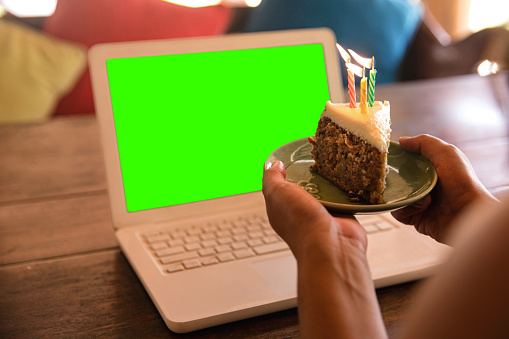 Close-up shot of unrecognizable woman holding a slice of birthday cake with lighted candles in front of her laptop. She's celebrating through video conference. The laptop device screen is in chroma key.