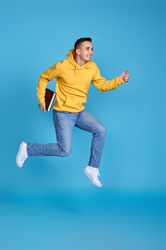 jumping very excited man with book in yellow sweatshirt and jeans isolated on blue background. Full length