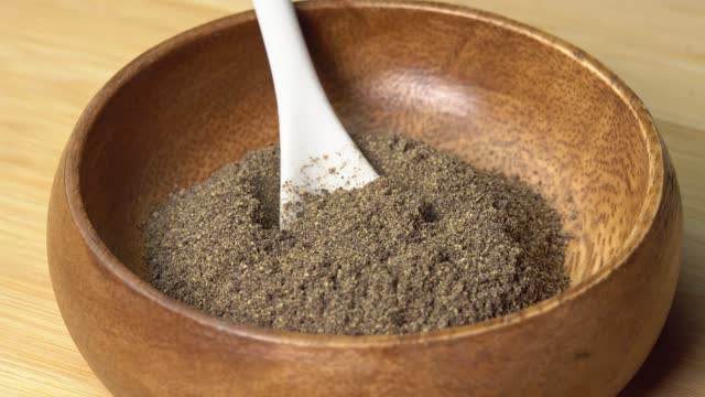 Ground black pepper spice in wooden bowl with spoon rotating on wooden board. Dry seasoning in spoon. Spices and herbs for cooking, pepper powder