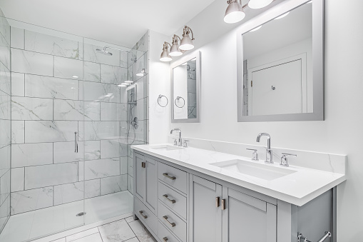 An elegant, remodeled bathroom with a grey vanity and bronze hardware. The shower has a large shower head and marble tiles and glass wall line the sides.