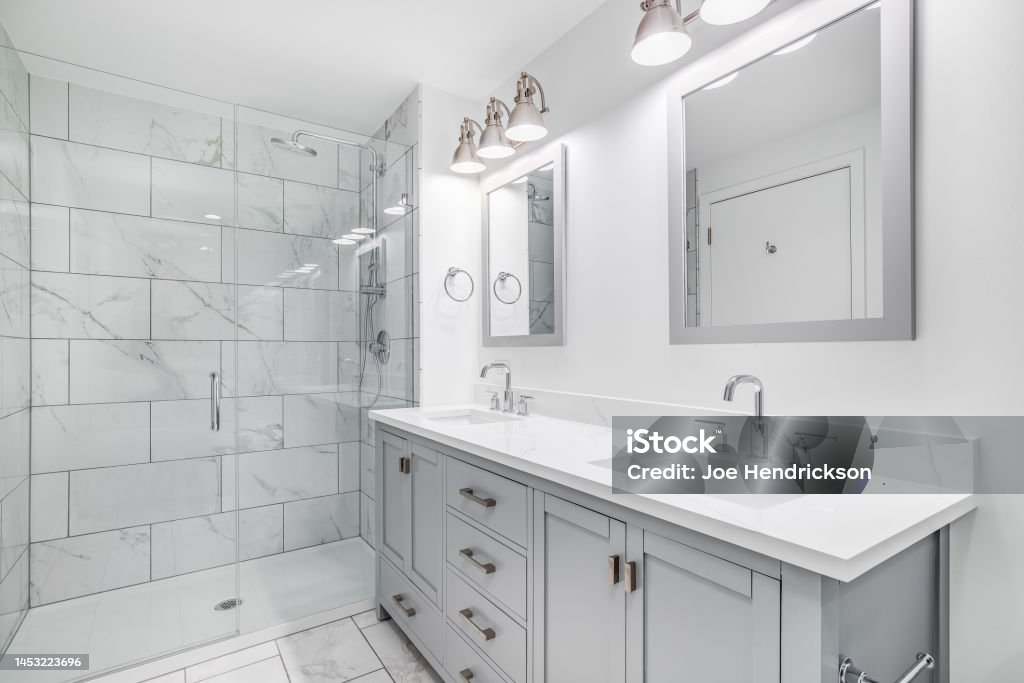 A bathroom with a grey cabinet and tiled shower. An elegant, remodeled bathroom with a grey vanity and bronze hardware. The shower has a large shower head and marble tiles and glass wall line the sides. Bathroom Stock Photo