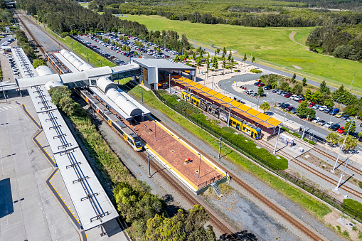 Helensvale, Australia - Nov 7, 2022: Aerial view of Helensvale public transport hub with railway station,  bus and Gold Coast Light Rail interchanges, car parks, green space. A Brisbane Airport-bound train is stationary in the nearest platform while across the tracks, a tram awaits passengers before departing for Broadbeach South. The bus station in the foreground has just the shadow of one bus visible. The station car parks are very busy.