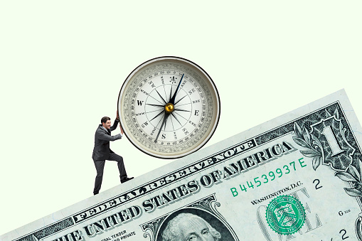 A man pushes a large compass on top of a large one dollar bill.