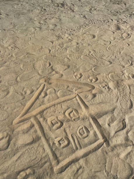 Stock photo showing close-up, elevated view of a house shape drawn in sand on a sunny beach with a stick. Real estate, vacation home and beach hut rental concept.