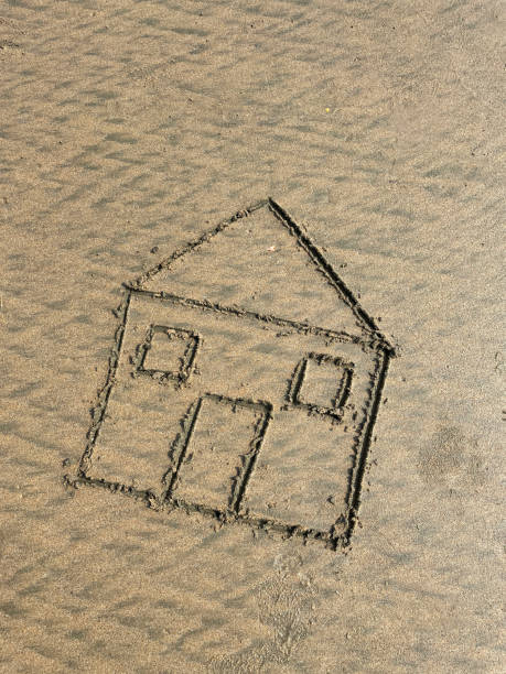 istock Image of house with two square windows and rectangular door drawn in damp sand with stick on a sunny beach, elevated view, copy space, real estate, holiday home and beach hut rental concept 1453244947