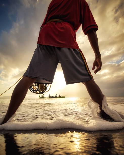 A fisherman reeling in his morning catch onto shore as a tradional fishing boat passes by in the horizon.\n\nLocation : Santen Island, Banyuwangi - Indonesia.