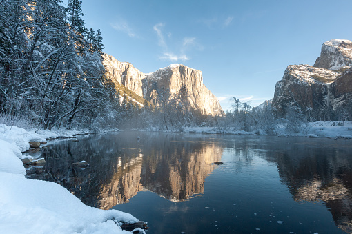 The rocky surface of the Sierra Nevada mountains, reflecting early morning sunlight, being reflected in Merced river, on an early winter, snow-covered landscape in Yosemite national park.