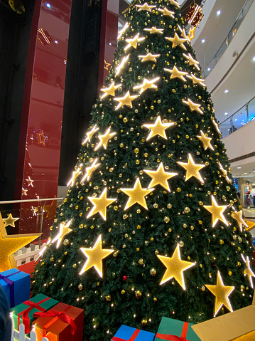 photo showing close-up view of outdoor, artificial Christmas tree covered in stars and gold baubles, with presents underneath, as part of a festive public seasonal display at a shopping mall.