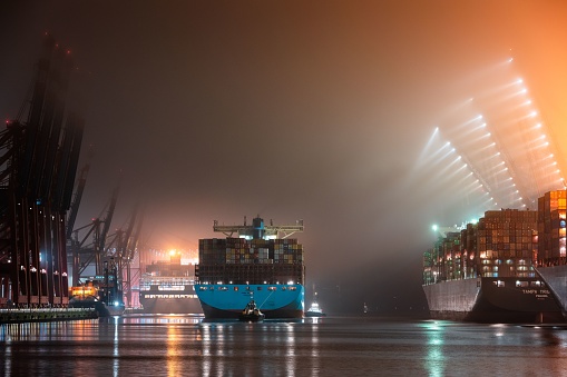 Hamburg, Germany – May 31, 2022: A nighttime shot of container ships and tugboats in the port of Hamburg in a foggy atmosphere