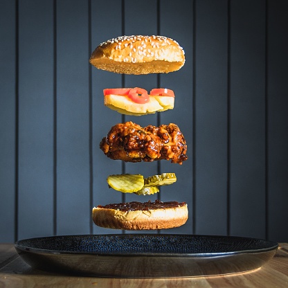 A vertical shot of a spicy chicken burger with gherkins, pineapple, and chili falling in stacks on a plate