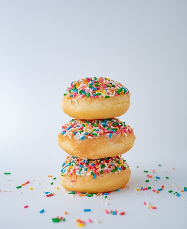 A vertical closeup of sprinkled donuts stacked on top of each other against the white background.