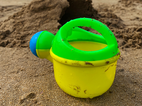 Close-up image of yellow, green and blue plastic toy watering can on beach, child's plaything on damp sand, focus on foreground