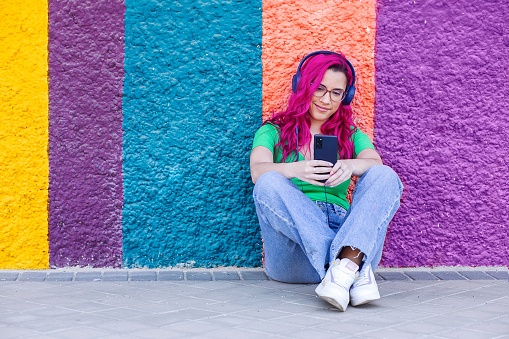 A beautiful young girl with pink hair and headphones sitting against a colorful textured wall