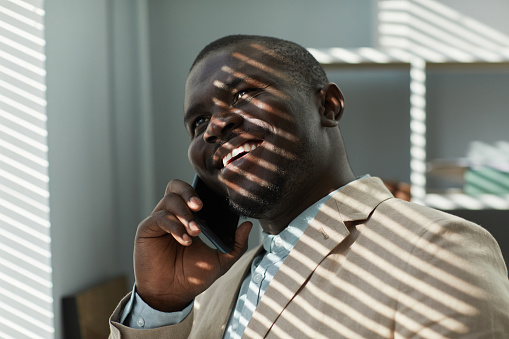 Close up portrait of smiling black businessman speaking on phone in office with sunlight rays