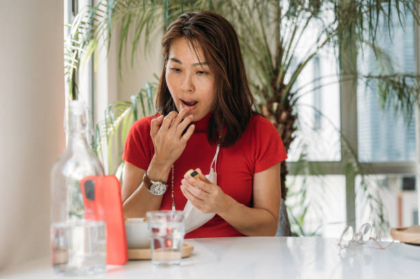 An Asian woman applying lip balm and using smart phone as a mirror at co working space stock photo