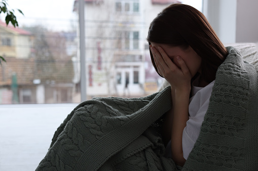 Sad young woman wrapped in warm plaid crying at home, space for text. Loneliness concept