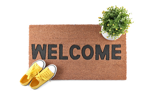 Stylish door mat with word Welcome, houseplant and shoes on white background, top view