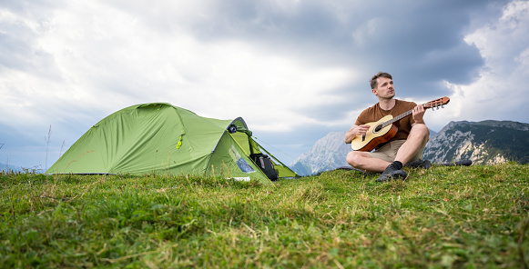 Man sitting in front of tent and playing guitar while camping in mountains. Healthy lifestyle and nature concept.