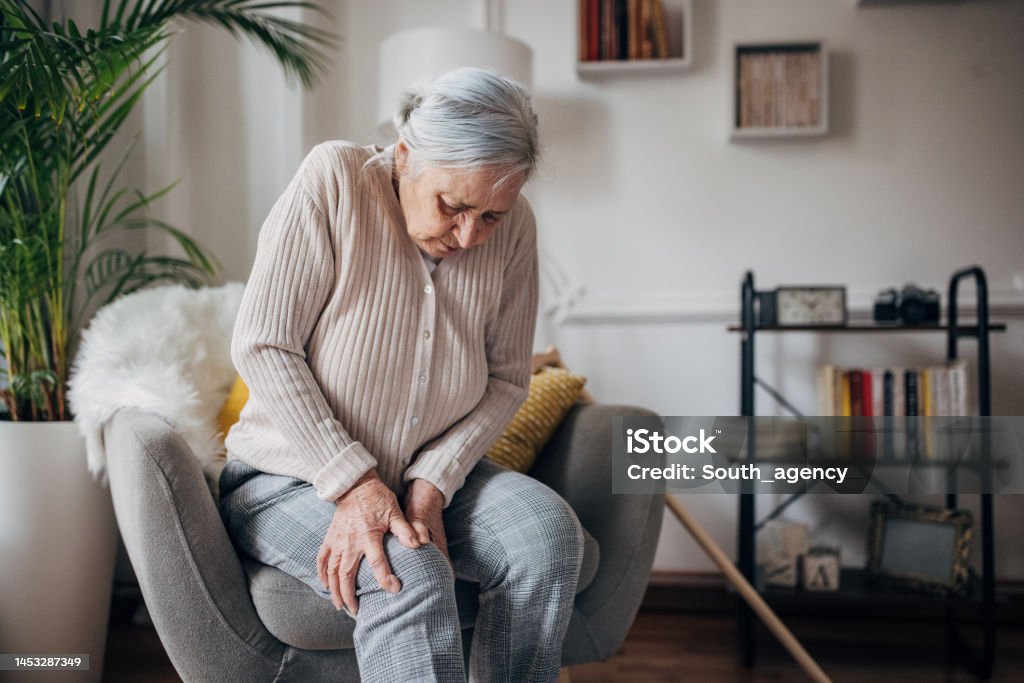 Old woman has knee pain One woman, old woman having back knee while sitting in armchair at home. Pain Stock Photo
