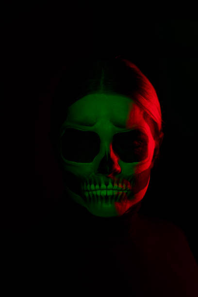 A woman with a painted skull face against a black wall. The woman is dressed in black and is illuminated by red & green light.