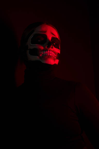 A woman with a painted skull face against a black wall. The woman is dressed in black and is illuminated by red light.