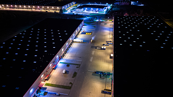 Aerial view of goods warehouse night. Logistics delivery; center in industrial city zone from above. Aerial view of trucks loading at logistic center. View from drone.