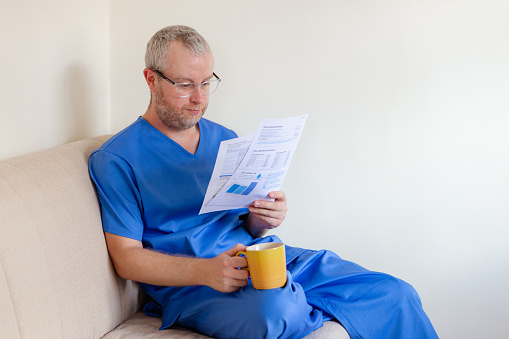 A male nurse in his 30s checks his energy bills at home. He is still wearing scrubs as he has just come home from work. The man has a worried expression as he examines the paper documents.