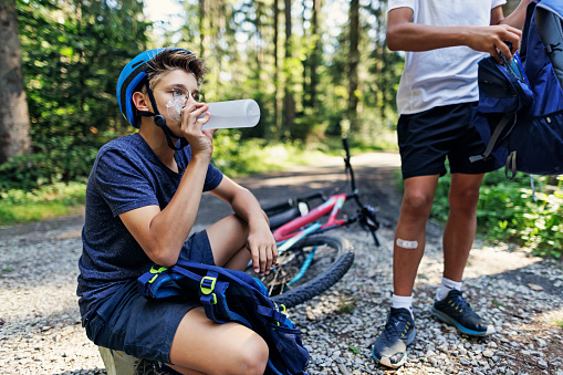 Mother and three teenage kids are enjoying a bike trip together in a forest. Teenage boy is drinking water from a reusable water bottle.\nCanon R5