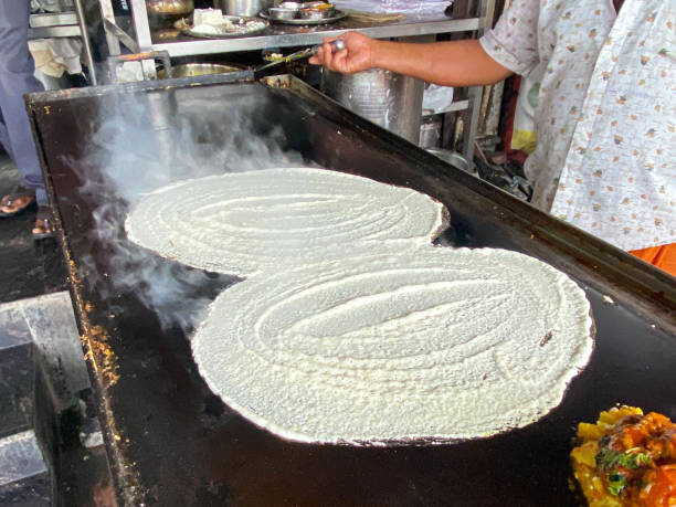 Image of dosa pancakes, traditional Indian street food being cooked on outdoor stove, unrecognisable chef holding metal spatula, smoking, hot oven top, elevated view, focus on foreground stock photo