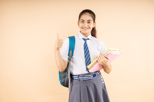 Happy Indian student schoolgirl do thumbs up wearing school uniform holding books and bag standing isolated over beige background, Studio shot, Education concept.