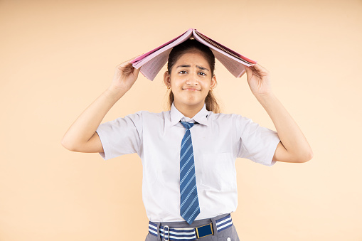 Unhappy Indian student schoolgirl wearing school uniform holding books on head standing isolated over beige background, Studio shot, Education concept.