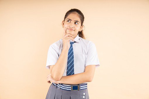 Modern Indian student schoolgirl wearing school uniform thinking expression standing isolated on beige background, Studio shot, closeup, Education concept.