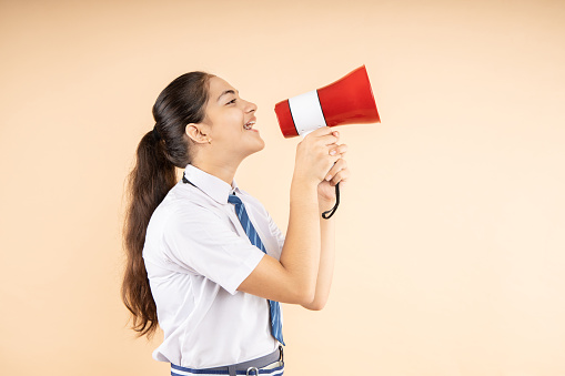 Happy Excited Indian student schoolgirl wearing school uniform hold screaming in megaphone or loud speaker isolated over beige background, Copy Space,Studio shot, Education concept.