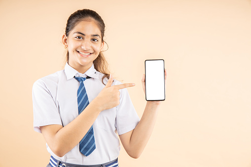 Happy Indian student schoolgirl wearing school uniform holding smart phone and pointing, blank screen to put advertisement isolated over beige background, Studio shot, closeup, Education concept.