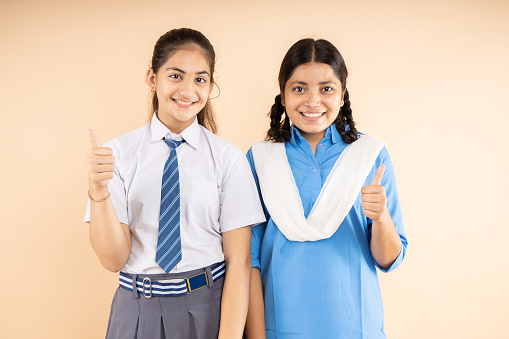 Happy Rural and Modern Indian student schoolgirls wearing school uniform standing together do thumbs up isolated over beige background, okay sign, all the best, Studio shot, Education concept.