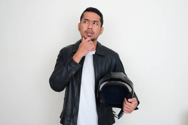 Pria Asia dewasa yang mengenakan jaket kulit memegang helm sepeda motor menunjukkan ekspresi berpikir foto stok