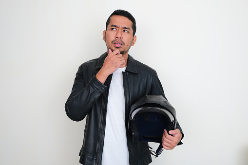 Adult Asian man wearing leather jacket holding motorcycle helmet showing thinking expression