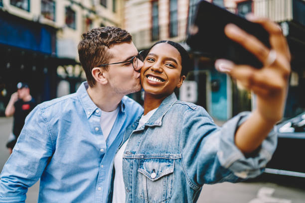 istock Caucasian hipster guy kissing happy dark skinned girlfriend while posing and making selfie during together trip for recreating, sincerely diverse marriage taking photo via mobile front camera 1453315882