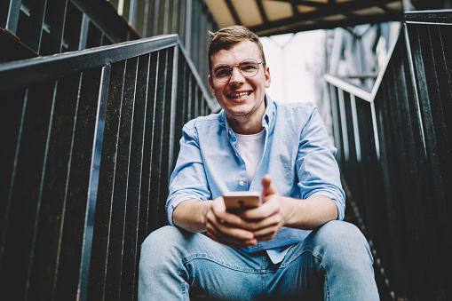 Joyful Caucasian male user holding cellphone gadget in hands and laughing at city urbanity, cheerful man in optical spectacles for provide eyes protection enjoying free time for networking outdoors