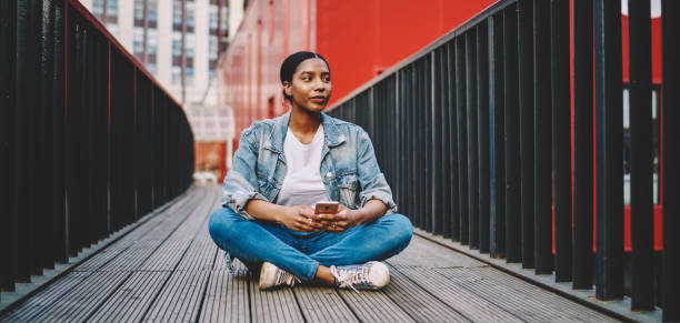 istock Pondering hipster girl taking rest at city urban setting holding modern cellular technology and looking away, thoughtful African American influence blogger 20s with smartphone thinking on content 1453315921
