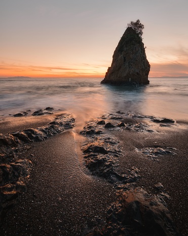 A vertical long exposure shot of sea waves breaking on the shore with a rock in the backdrop at sunset