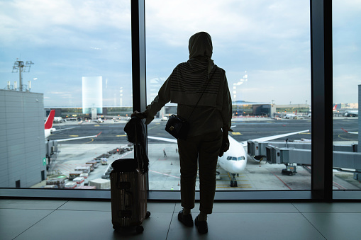 Travel tourist standing with luggage watching sunset at airport window. Woman looking at lounge looking at airplanes while waiting at boarding gate before departure.