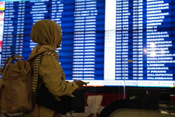 woman with luggage and smart phone checking flight schedule near departure board in airport.