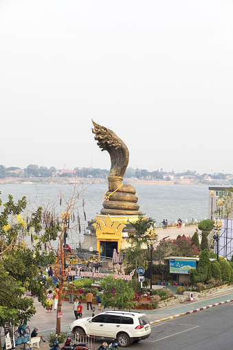 Morning high angle view of gold colored Naga statue at promenade of Nakhon Phanom in east Thailand in morning. A few people are praying and kneeling in front of statue which is local landmark. In background is Mekong river