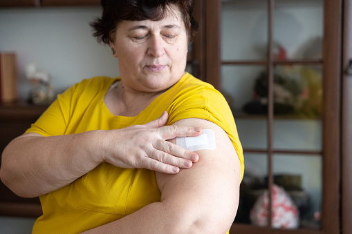 The woman applies one HRT patch to the skin into the outside of upper arm. The woman is standing in the living room. She is wearing a yellow T-shirt.